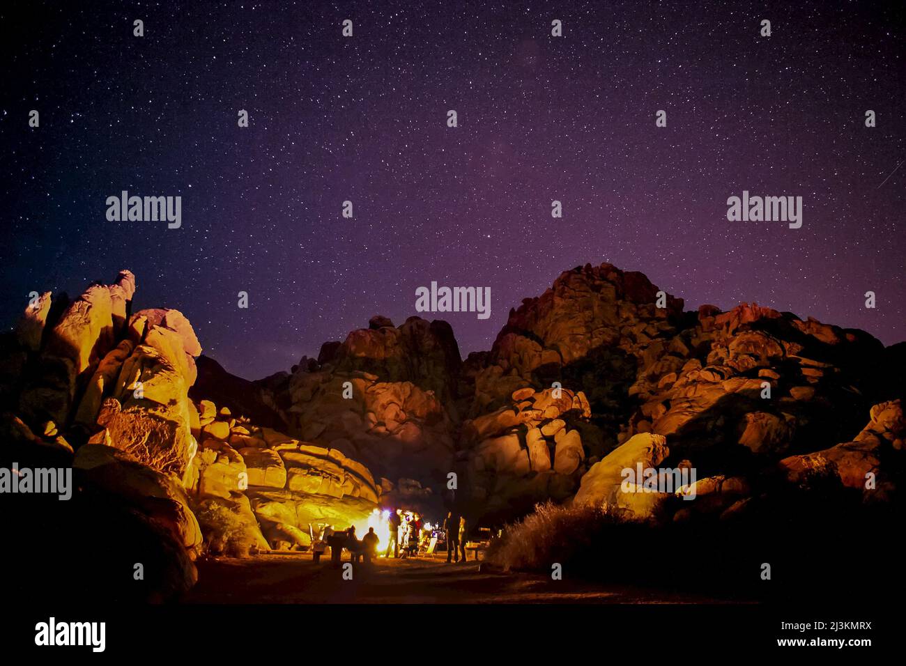 A group by a campfire that lights up granite rocks under a starlit sky. Stock Photo