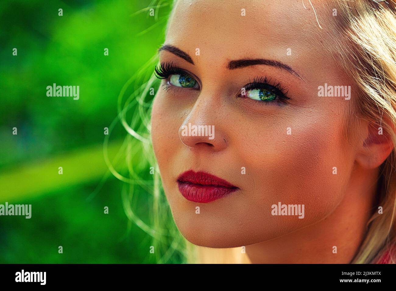 Attractive young blond woman wearing makeup turning to look quietly at camera with lovely blue eyes in a close up cropped face portrait outdoors again Stock Photo