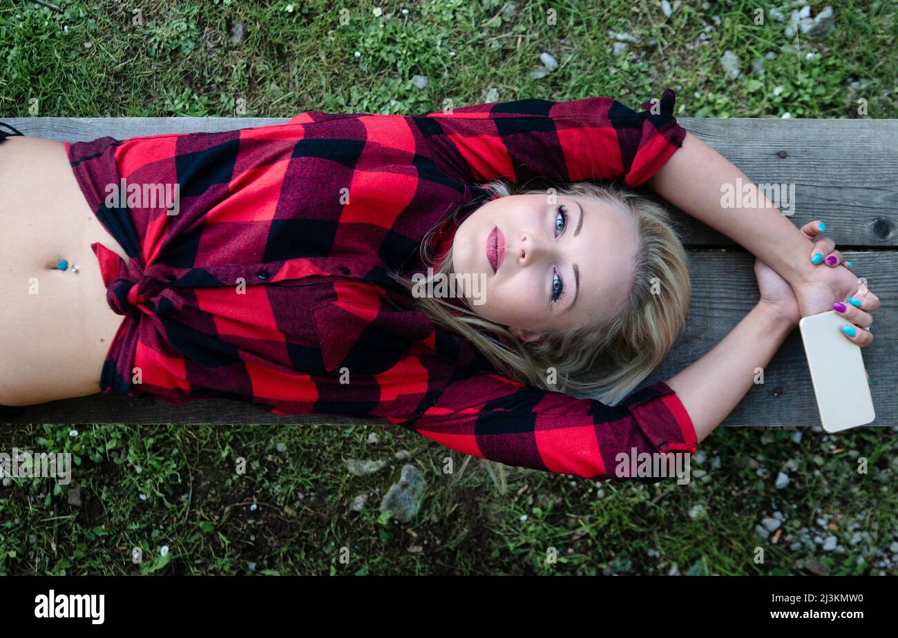 Trendy young woman with belly piercings lying stretched out on a bench over green grass looking up at the camera with a thoughtful expression Stock Photo