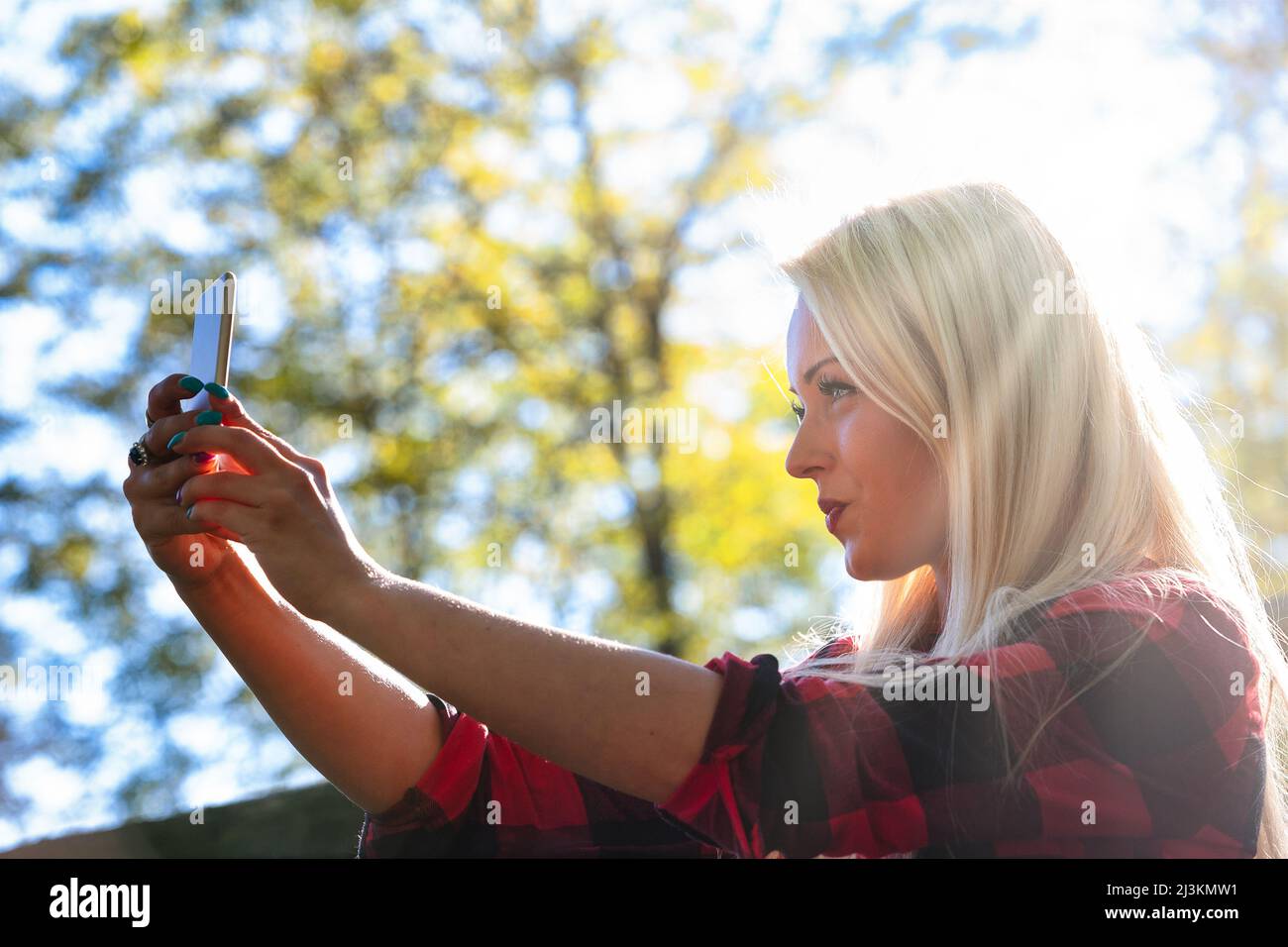 High key portrait of a young woman taking a backlit selfie on her mobile phone outdoors against leafy green trees in a low angle view Stock Photo