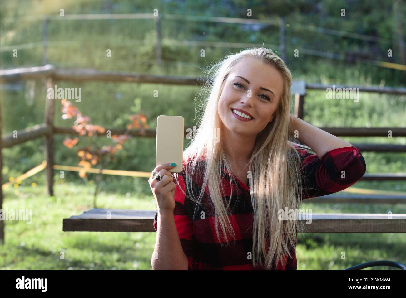 Attractive young blond woman posing holding up a mobile phone with her hand to her hair and a lovely warm smile outdoors on a bench in a garden Stock Photo