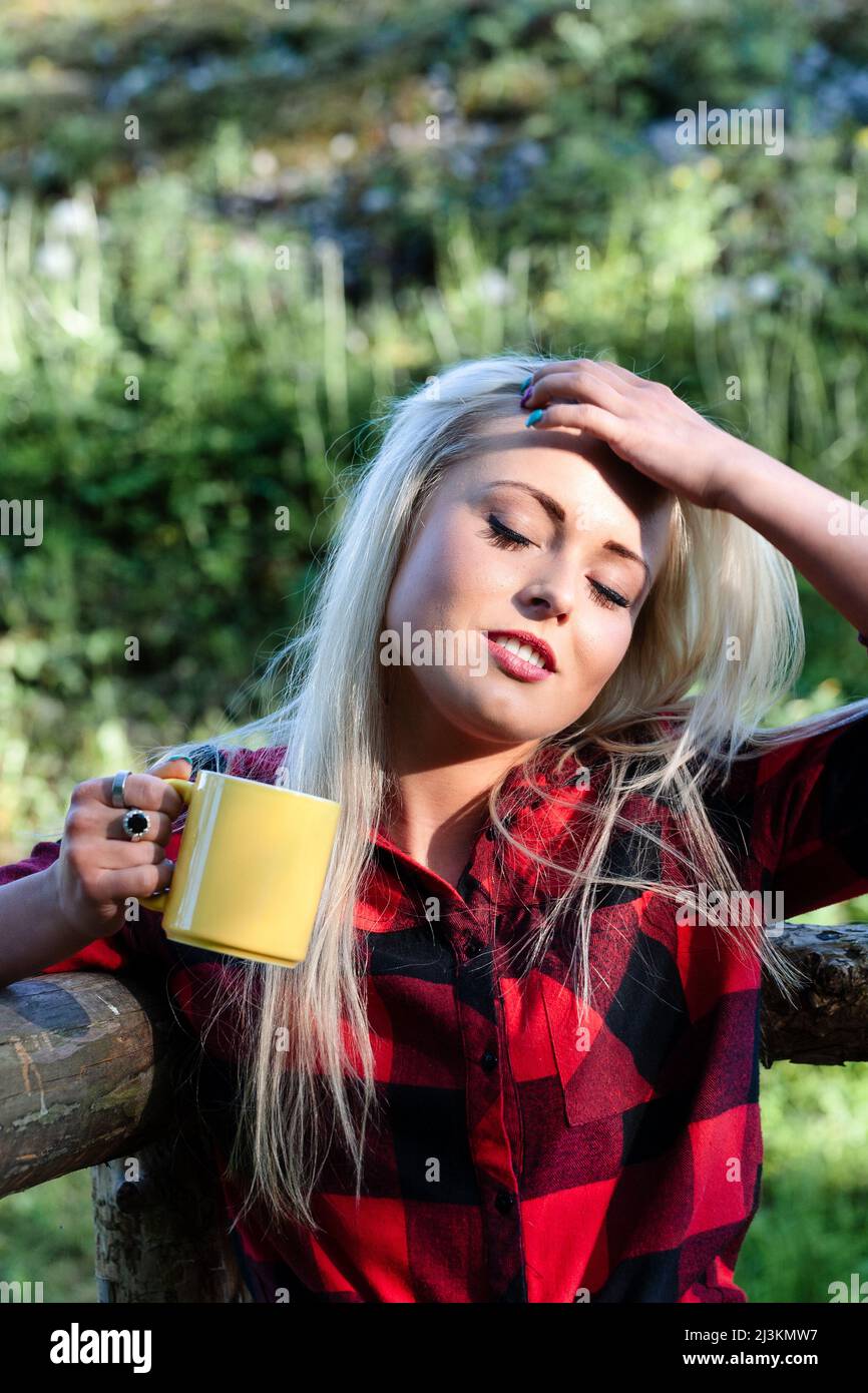 Young woman enjoying the warm sunshine outdoors standing holding a mug of coffee in one hand with the other to her forehead with closed eys and face t Stock Photo