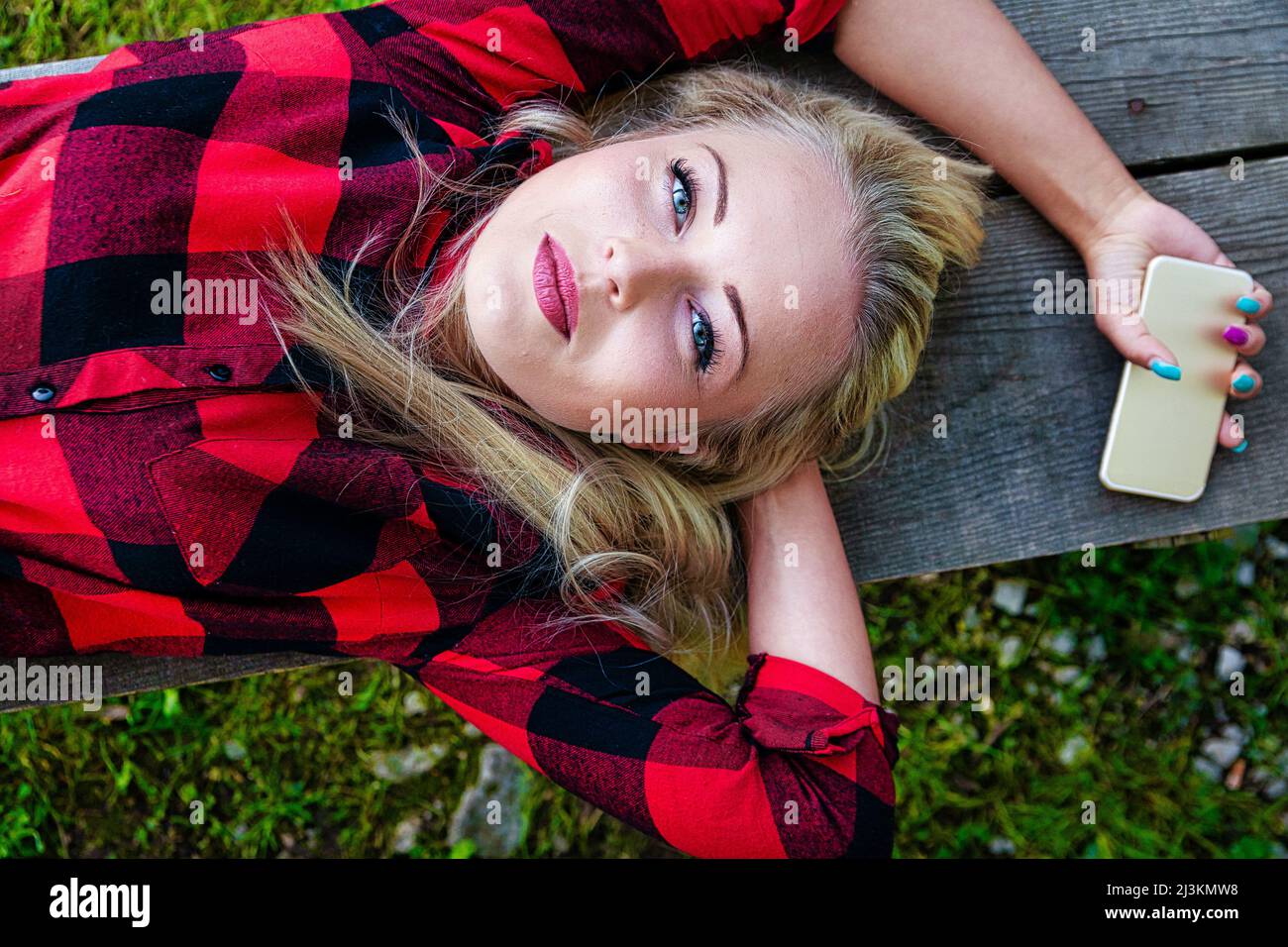 Pensive young blond woman with gorgeous blue eyes lying stretched out on a park bench looking up at the camera with a quiet calm expression Stock Photo