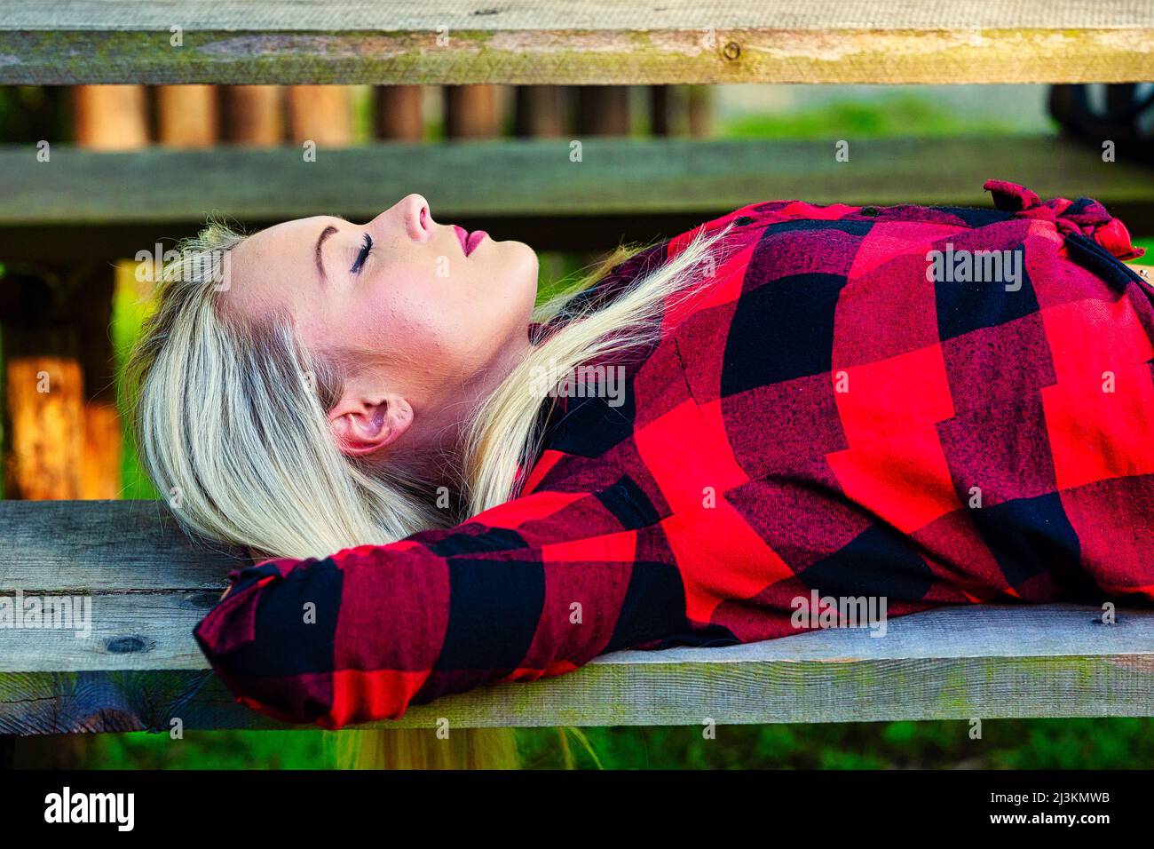 Serene young woman enjoying a sleep or nap on a park bench lying back with hands behind her head in a colorful red shirt in a close up side view Stock Photo