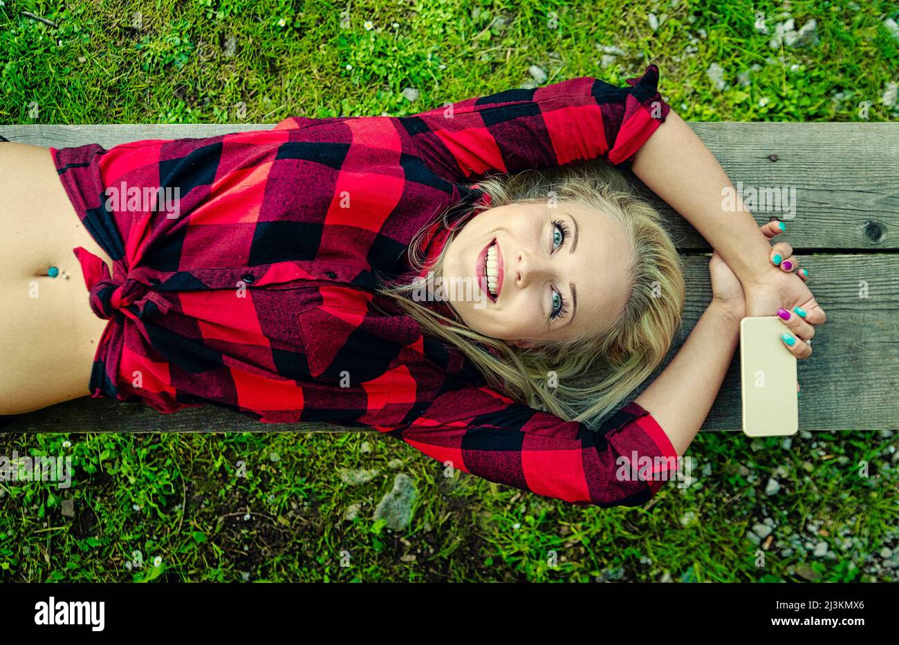 Happy pretty young blond woman daydreaming with blissful smile and beatific expression as she lies back on a wooden bench over grass Stock Photo