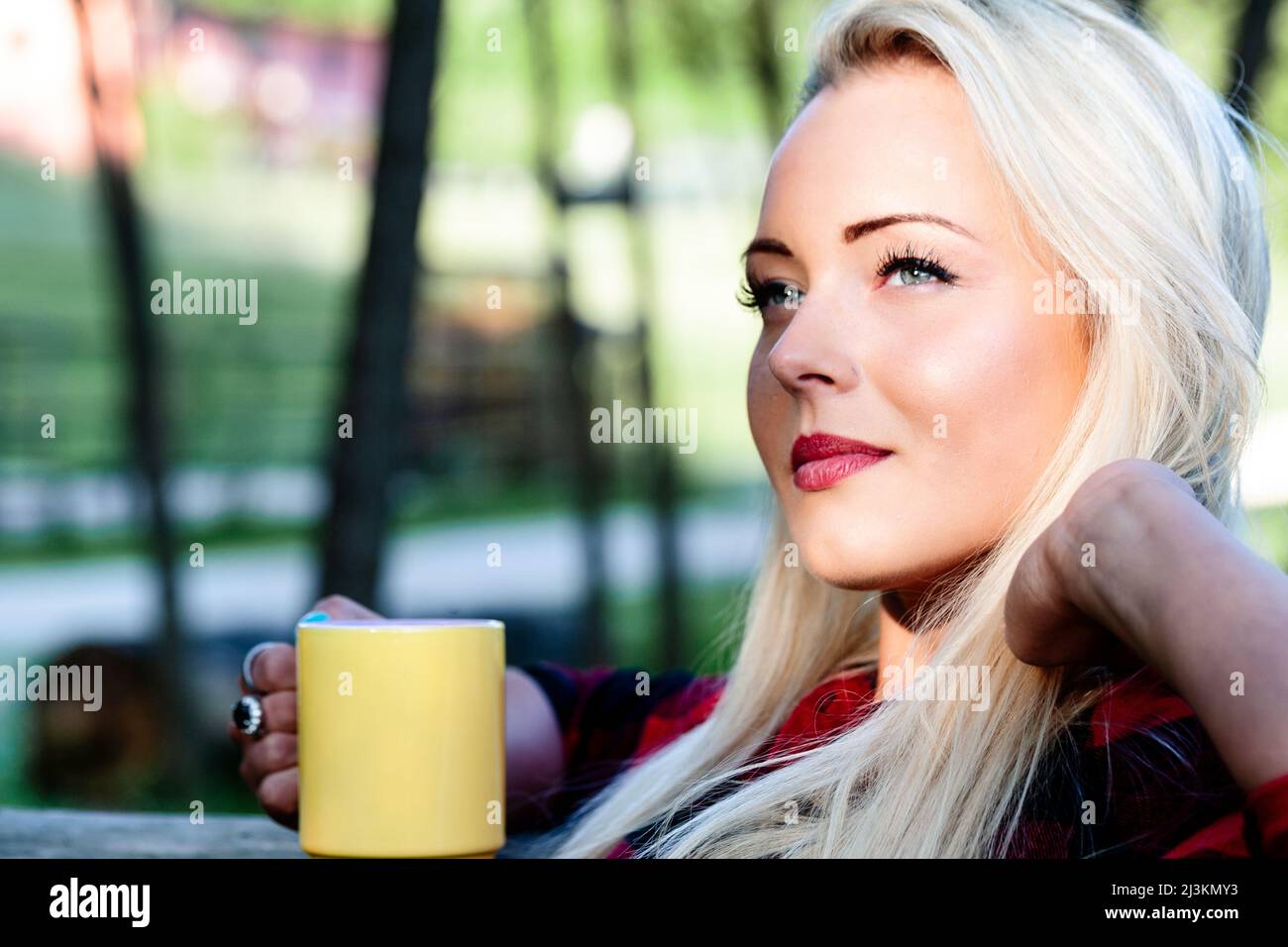 Beautiful young blond woman sitting daydreaming while enjoying a mug of hot coffee looking up at the sky with a serene pensive expression reliving her Stock Photo