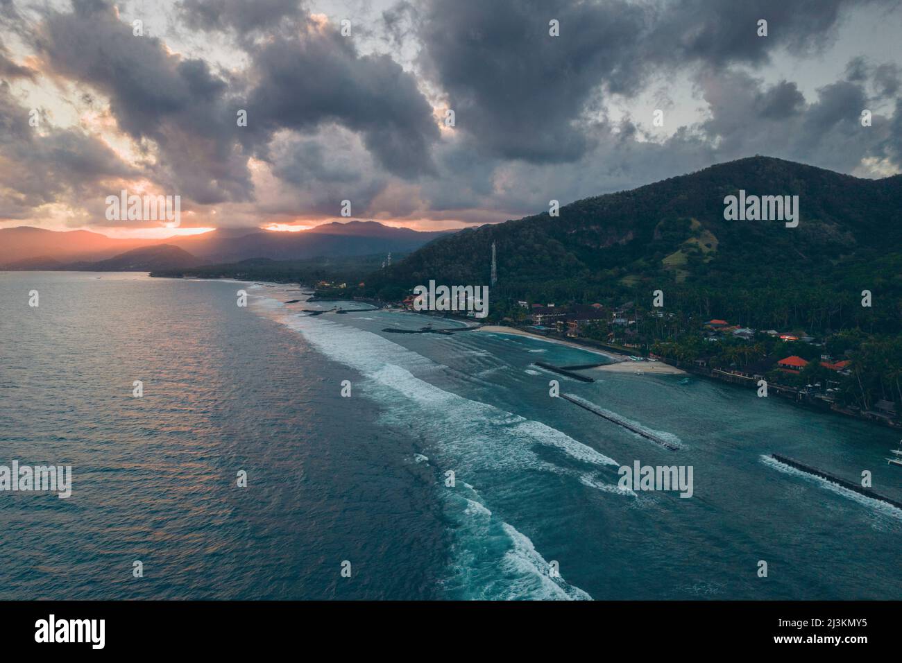 Aerial view of the docks and breakwaters along the shore of Candidasa Beach with sunlight streaming across the mountaintops under a grey cloudy sky... Stock Photo