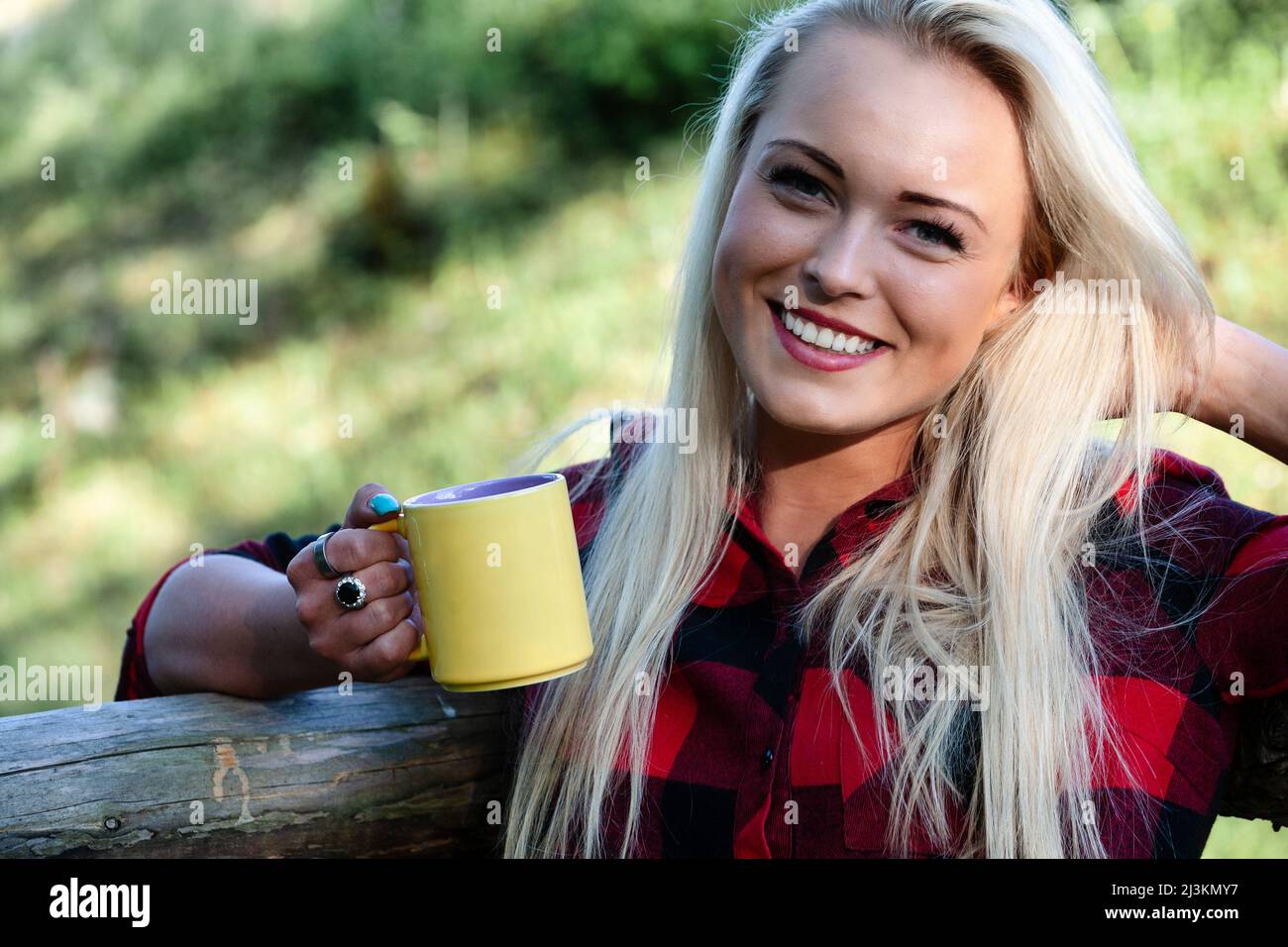 Beautiful happy smiling blond woman drinking a mug of hot coffee as she relaxes outdoors leaning on a wooden railing Stock Photo