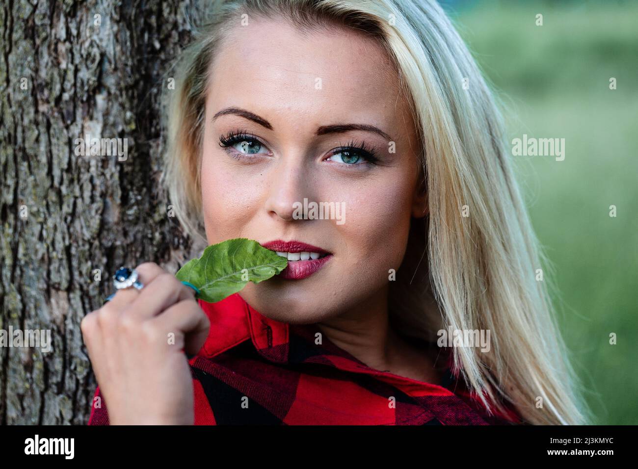 Pensive sensual young blond woman with green leaf to her lips relaxing outdoors looking at the camera with her lovely blue eyes Stock Photo