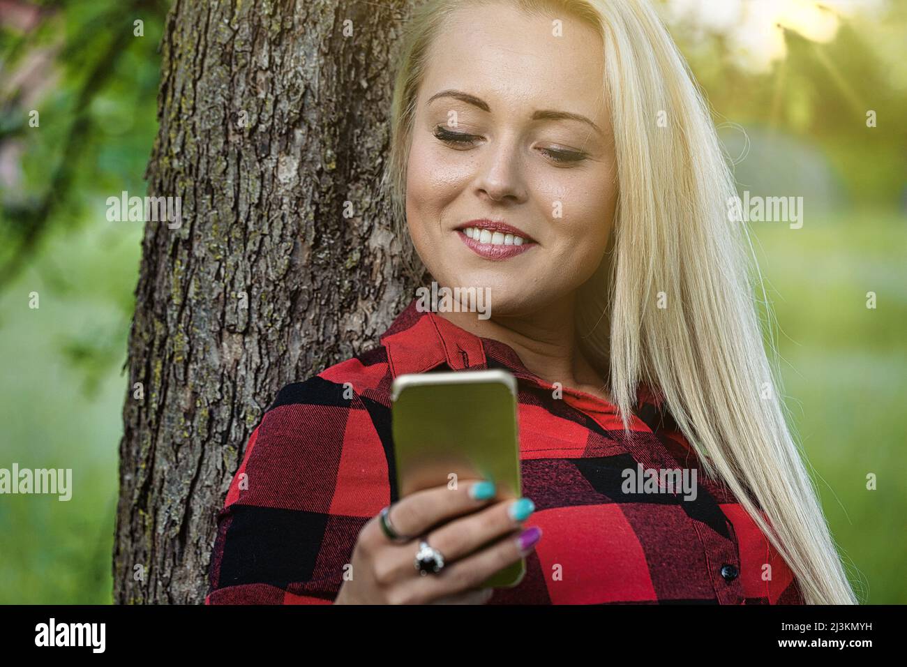 Young woman leaning against a tree trunk in a park with a mobile phone in her hands smiling happily backlit by the warm glow of the sun through trees Stock Photo
