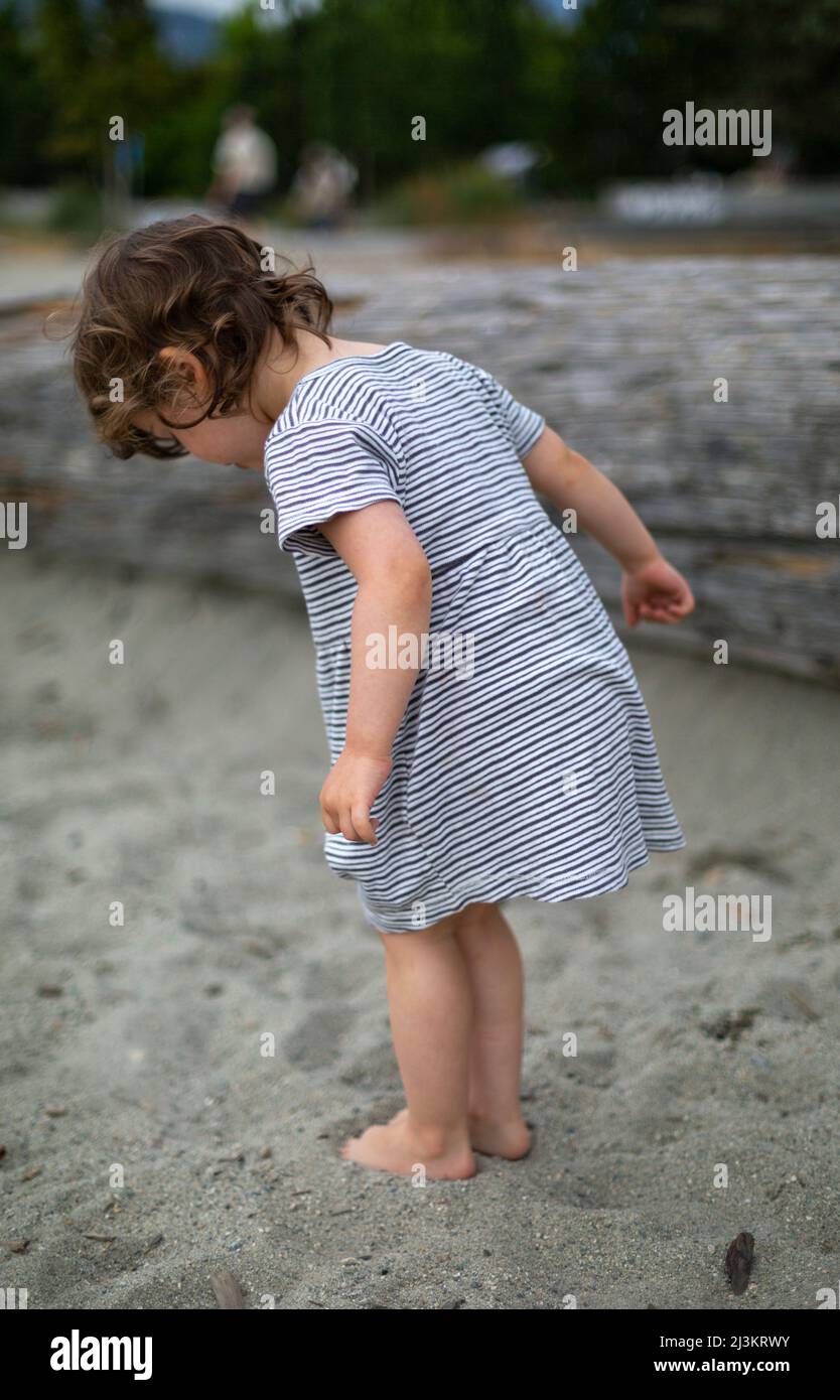 Preschooler girl standing on a beach digging her toes into the sand, Ambleside Beach in West Vancouver, BC, Canada Stock Photo