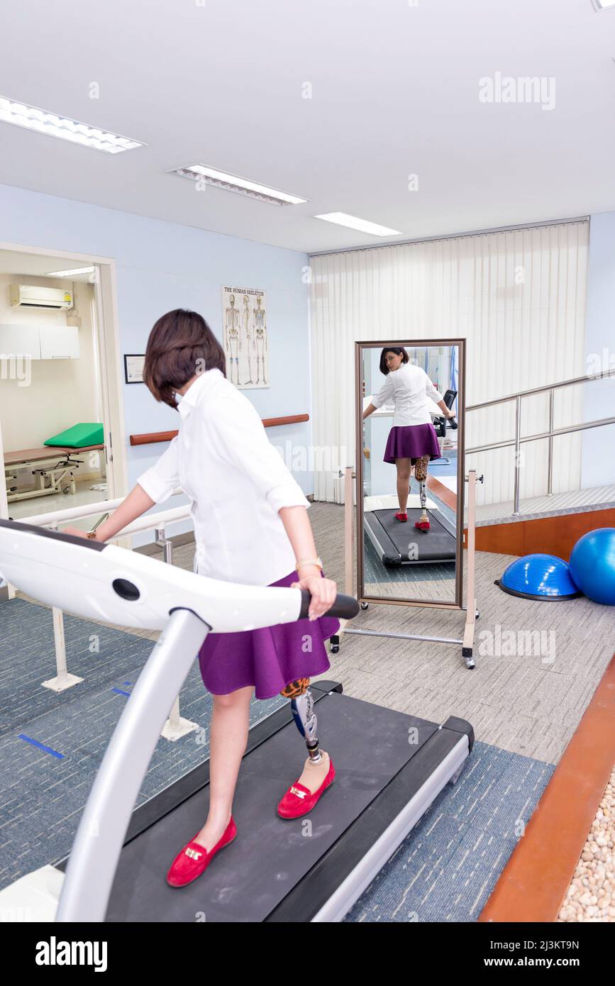 Young woman with leg prosthesis in therapy, walking on a treadmill and looking in the mirror at her reflection as she walks; Bangkok, Thailand Stock Photo