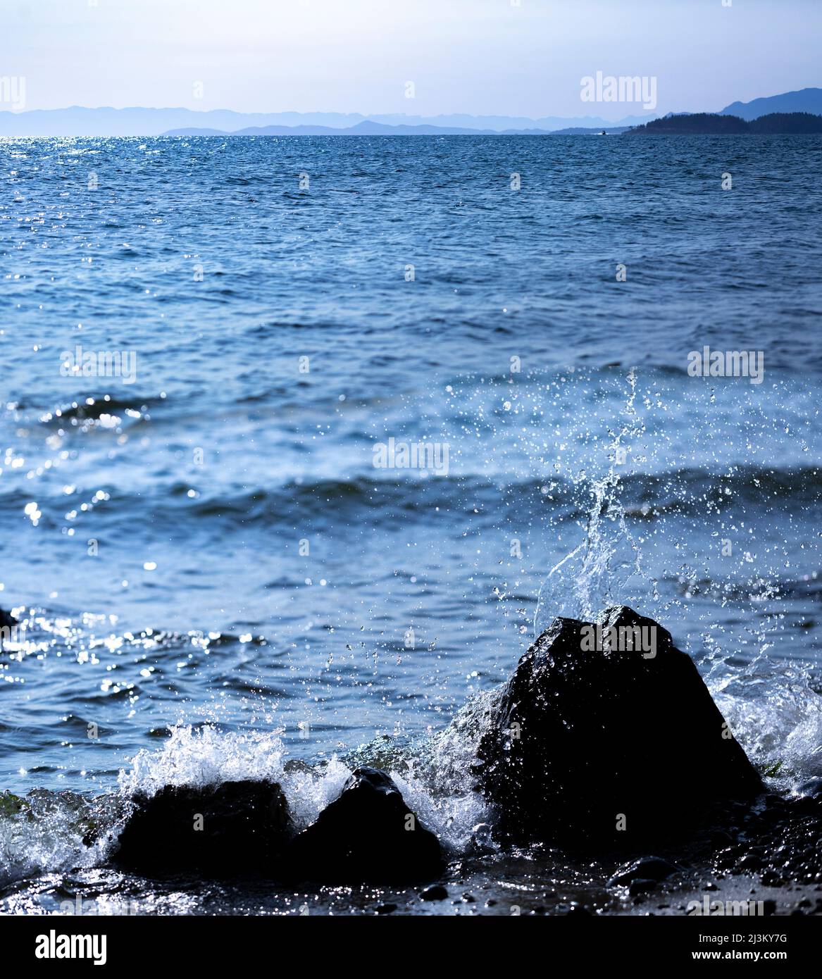 Splashing surf against rocks in the foreground with a view of the vast coastline along the Sunshine Coast; British Columbia, Canada Stock Photo