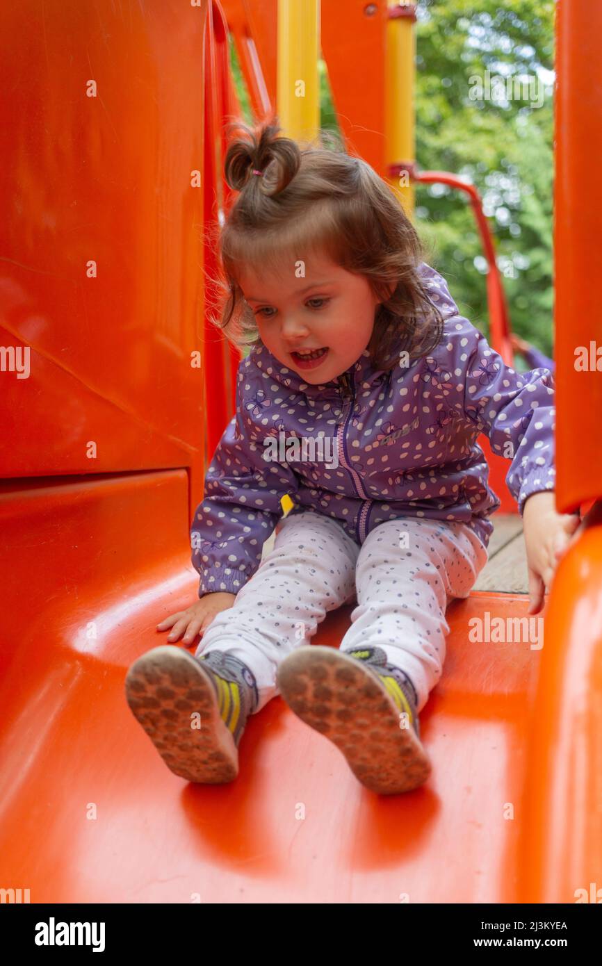 Preschool girl at a playground sitting at the top of a red slide; North Vancouver, British Columbia, Canada Stock Photo
