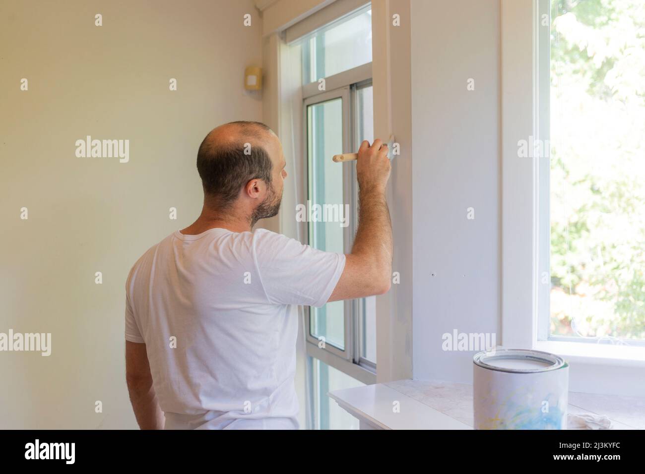 Man stands to paint a wall and trim with a paintbrush at home; North Vancouver, British Columbia, Canada Stock Photo
