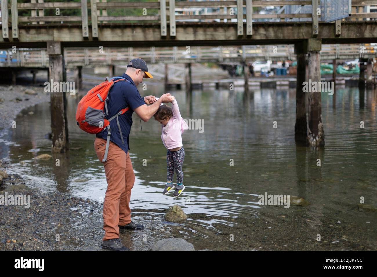 Father and young daughter being playful at the water's edge of a beach at the harbour of Horseshoe Bay in BC, Canada Stock Photo