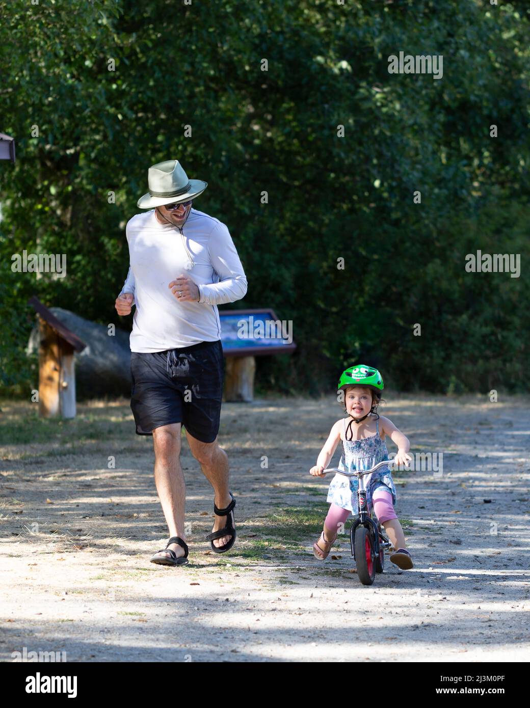 Father runs beside young daughter riding a glider bike; British Columbia, Canada Stock Photo
