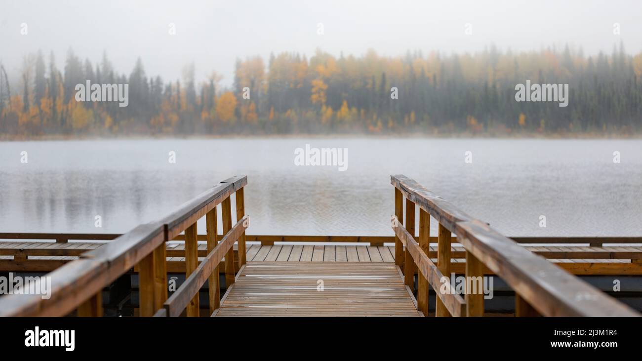 Dock on Hush Lake with fog blurring the autumn coloured forest along the shoreline; British Columbia, Canada Stock Photo