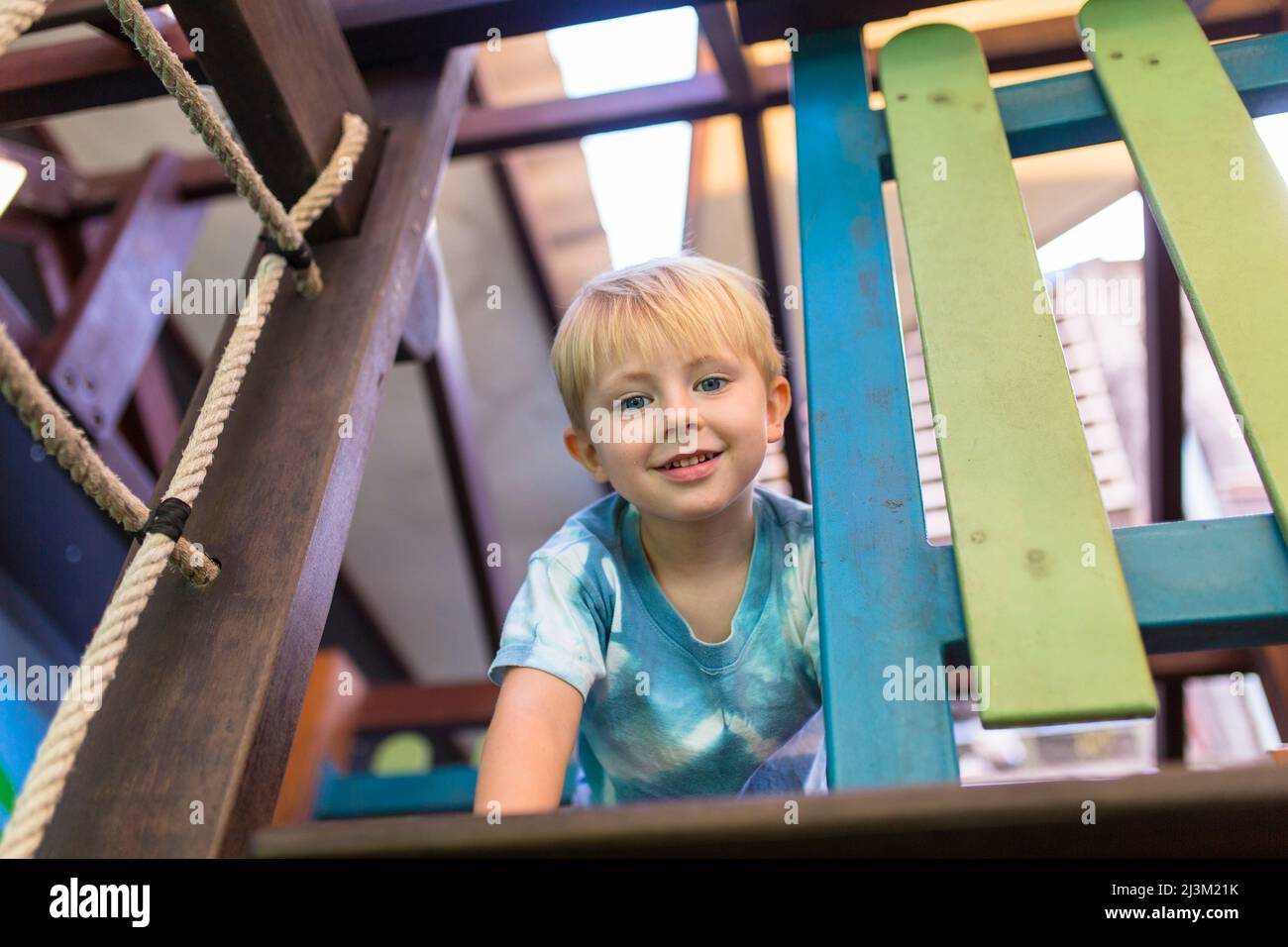 Young boy with blue eyes and blond hair looks down at the camera while climbing on a play structure in a park; Vientiane, Vientiane Prefecture, Laos Stock Photo