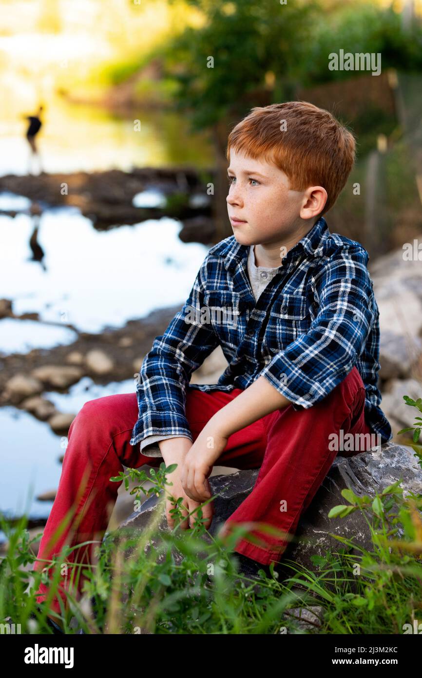 Outdoor portrait of a boy with red hair sitting beside a creek; Edmonton, Alberta, Canada Stock Photo