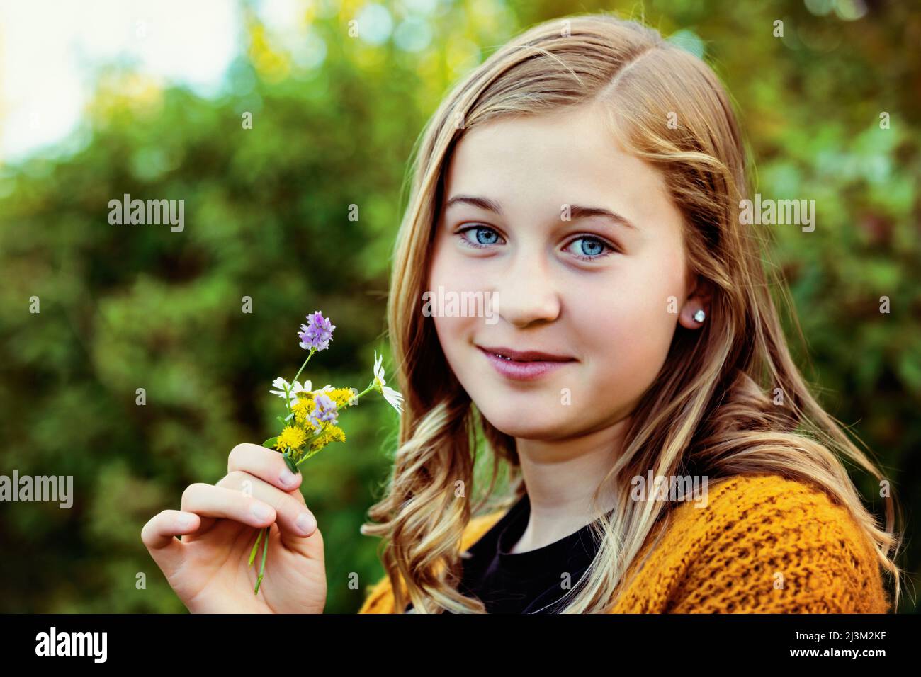 Teenage girl stands outdoors holding a small cluster of colourful wildflowers and looking at the camera; Edmonton, Alberta, Canada Stock Photo