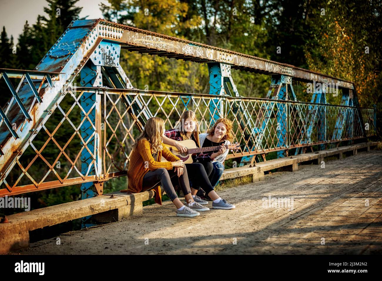 Mother and two sisters sit on a park bridge, one with an acoustic guitar, enjoying music together; Edmonton, Alberta, Canada Stock Photo