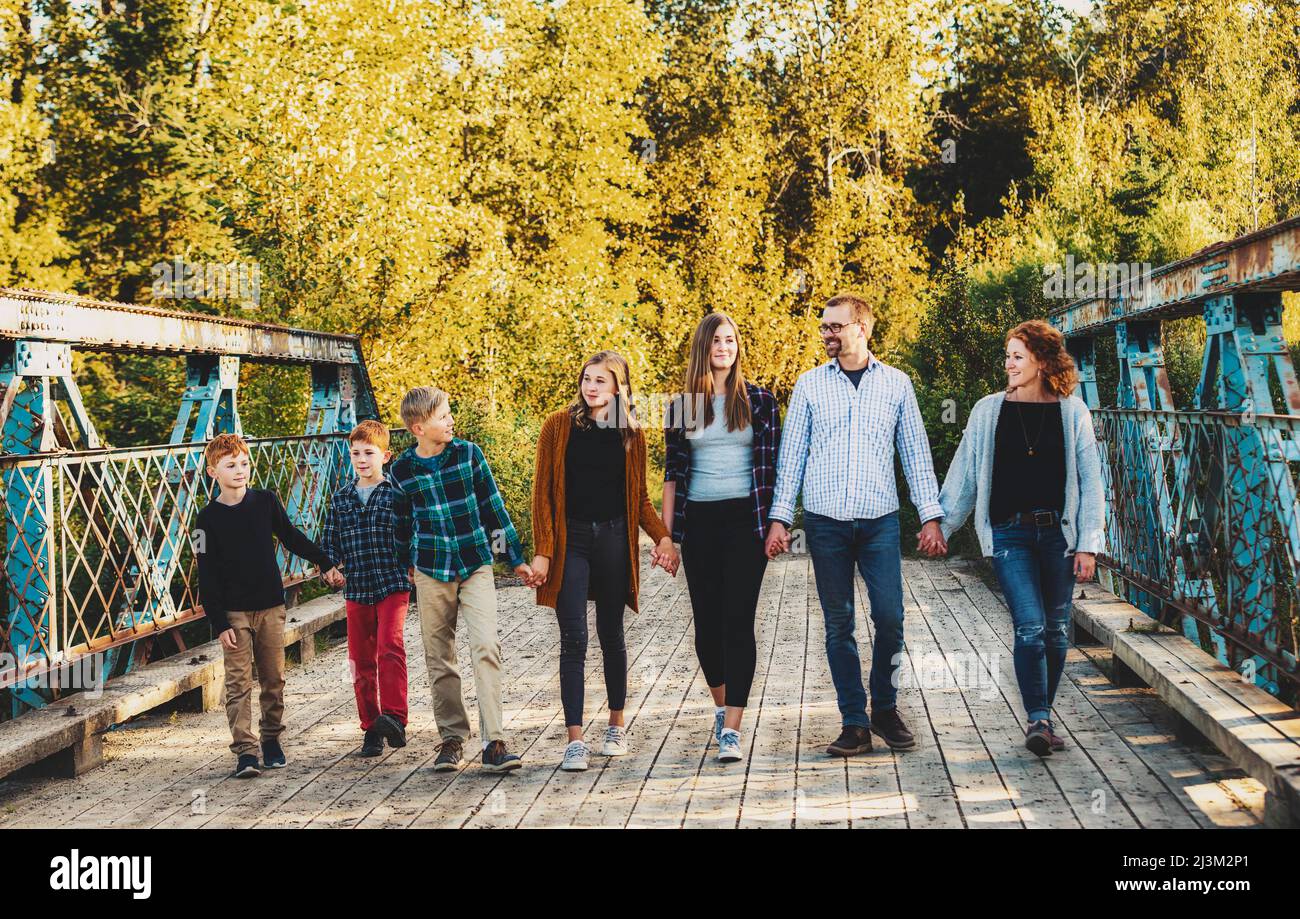 Family of seven walking in a row and holding hands as they walk across a bridge in a city park in autumn; Edmonton, Alberta, Canada Stock Photo