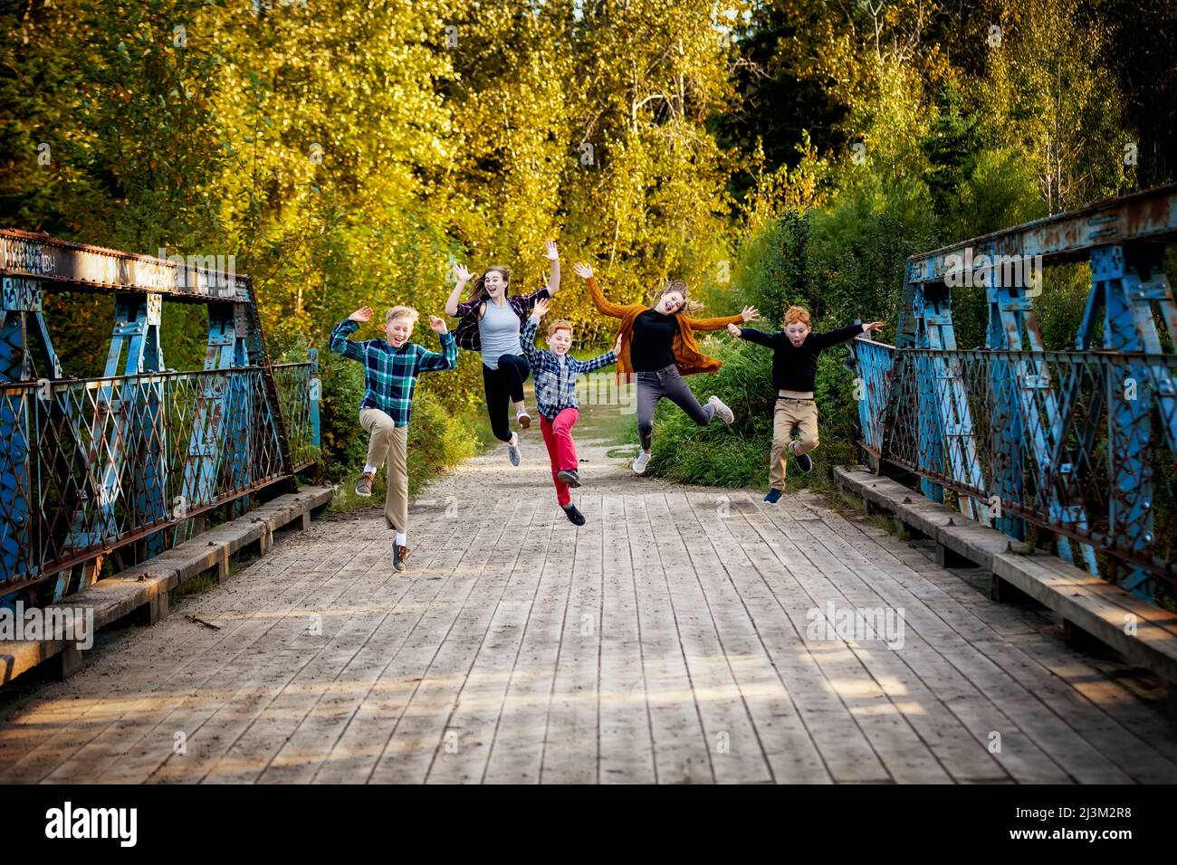 Five siblings in a row jump in mid-air making funny expressions on a park bridge; Edmonton, Alberta, Canada Stock Photo