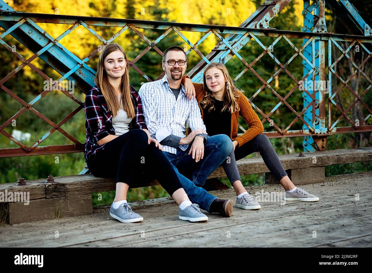 Outdoor portrait of a father with two teenage daughters sitting on a bridge in a city park in autumn; Edmonton, Alberta, Canada Stock Photo