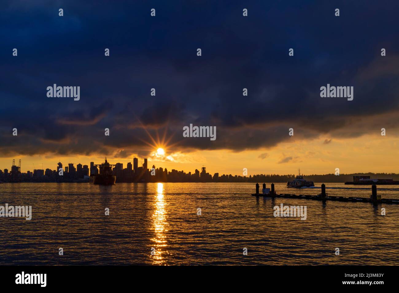 Golden sun setting behind the downtown Vancouver skyline and horizon with sunbeam reflecting on the water of Burrard Inlet Stock Photo