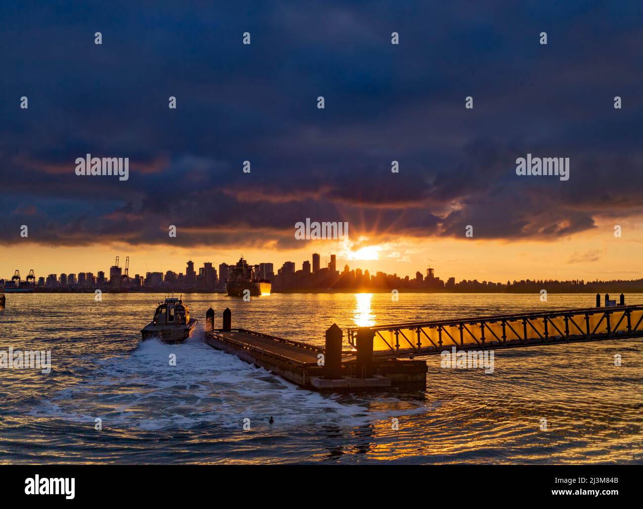 Speedboat leaves from the dock as the golden sun sets behind the downtown Vancouver skyline and horizon with sunbeam reflecting on the water of Bur... Stock Photo