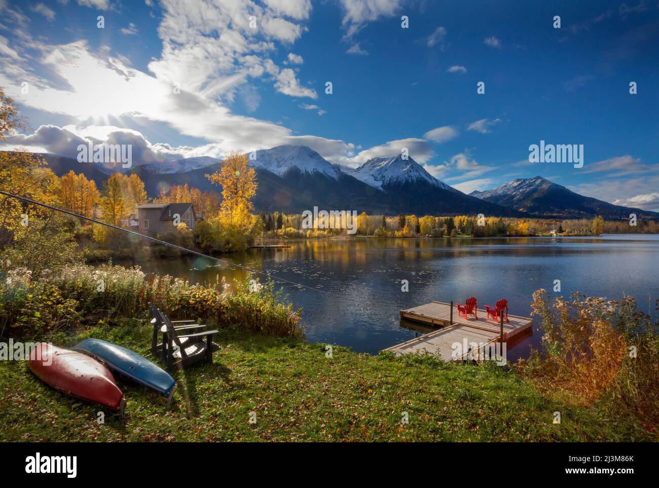 Two red chairs on a deck on tranquil Lake Kathlyn surrounded by autumn coloured trees and snow-capped Coast Mountains, viewed from Lakedrop Inn, Wa... Stock Photo