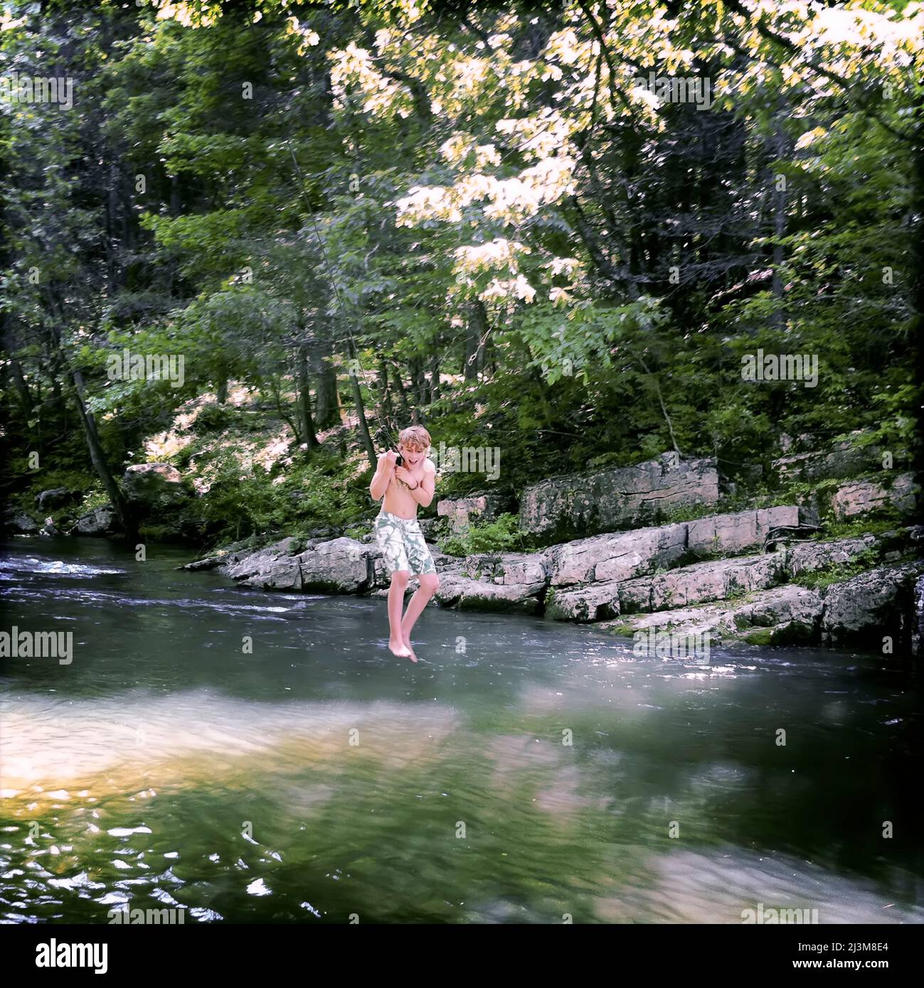 A boy swings out over a swimming hole on a rope swing on a summer day.; Highland County, Virginia, United States. Stock Photo