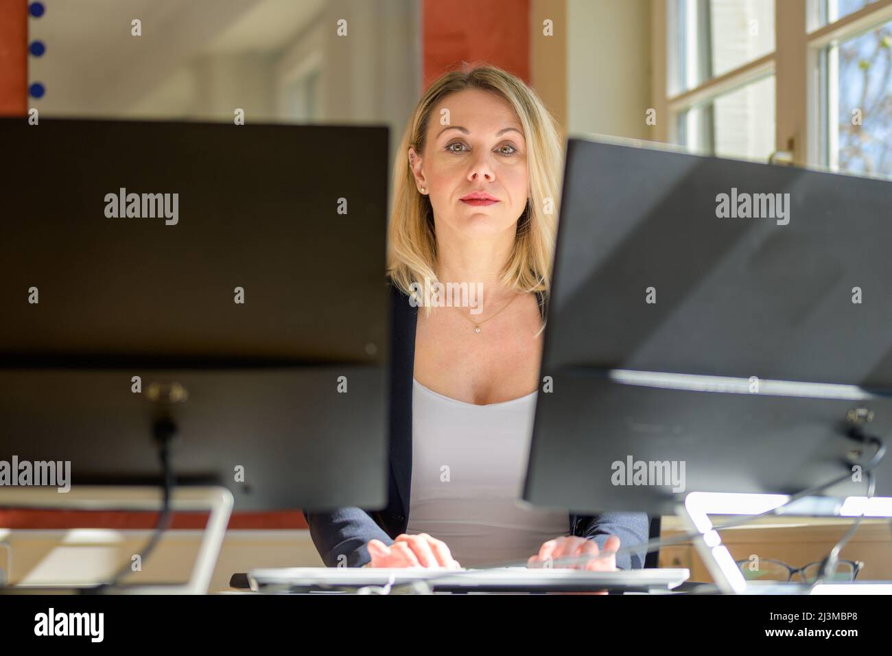Confident businesswoman or manageress looking at the camera with a thoughtful expression viewed between two computer monitors in an office Stock Photo