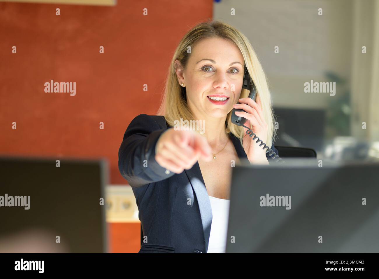 Portrait of business woman in formal clothing pointing towards camera while in a conversation over landline telephone in office Stock Photo