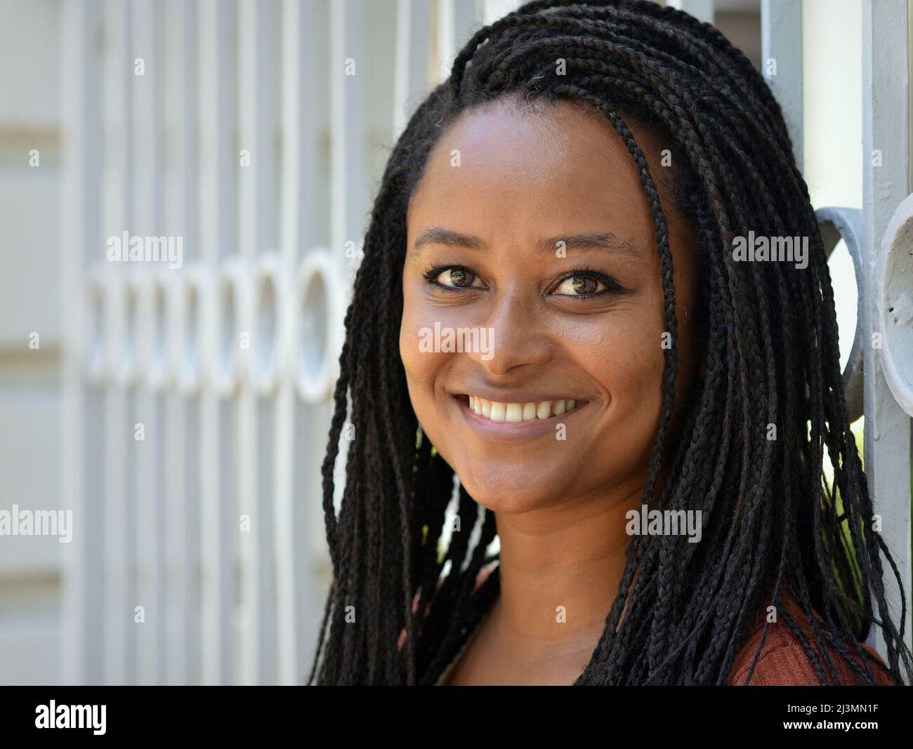 Cheerful positive beautiful young dark skinned African woman with long stylish Afro braids smiles for the viewer in front of a wrought iron fence. Stock Photo