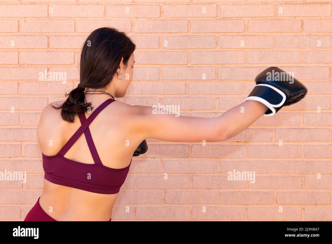 An unknown young Caucasian woman with boxing gloves is punching seen from the side and her back. The arm movement is captured in the photograph. The b Stock Photo
