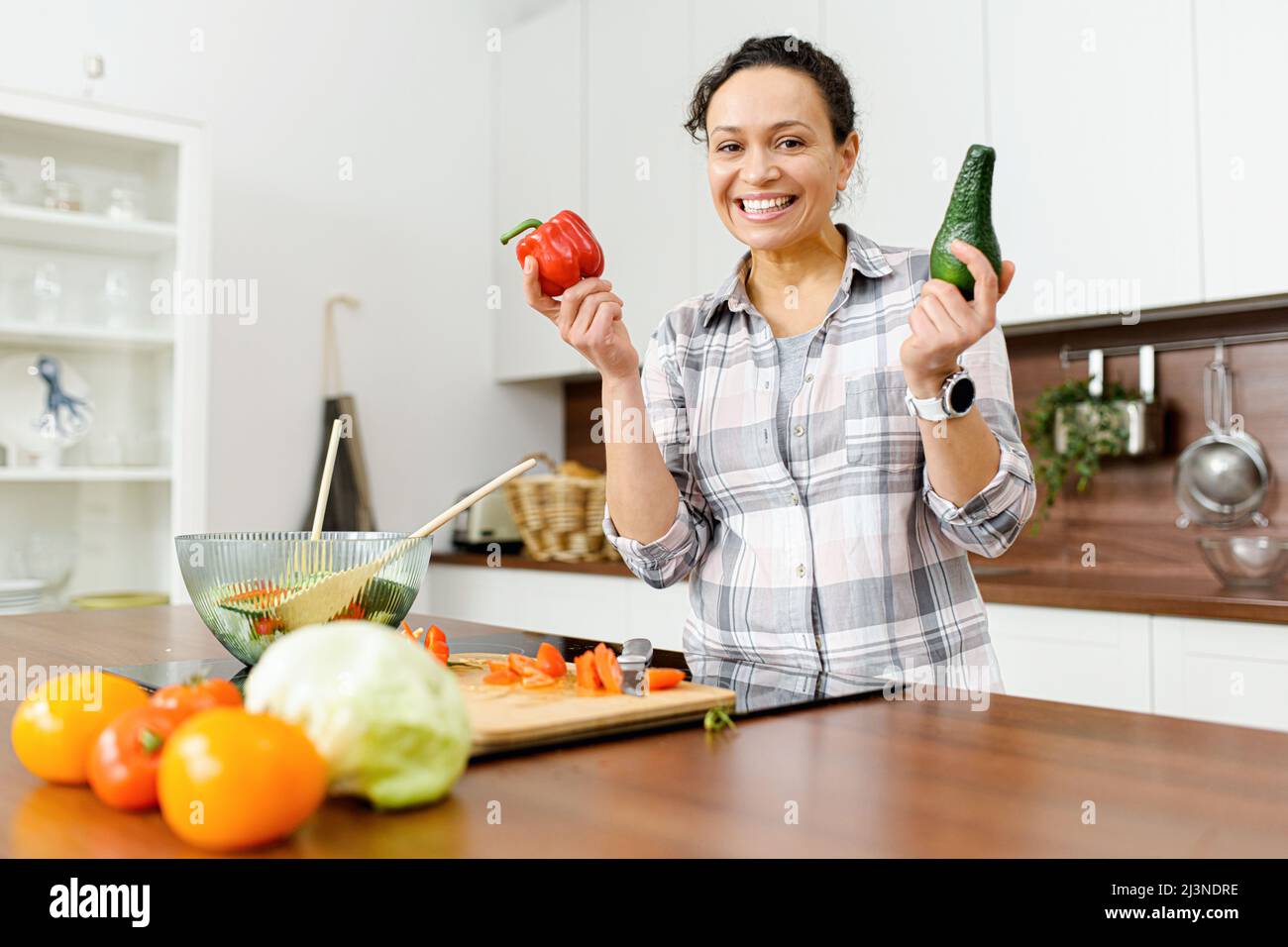 Smiling middle-aged woman going to cooking vegetable meals, standing in the modern kitchen, holding avocado and pepper in hands, looking at camera Stock Photo