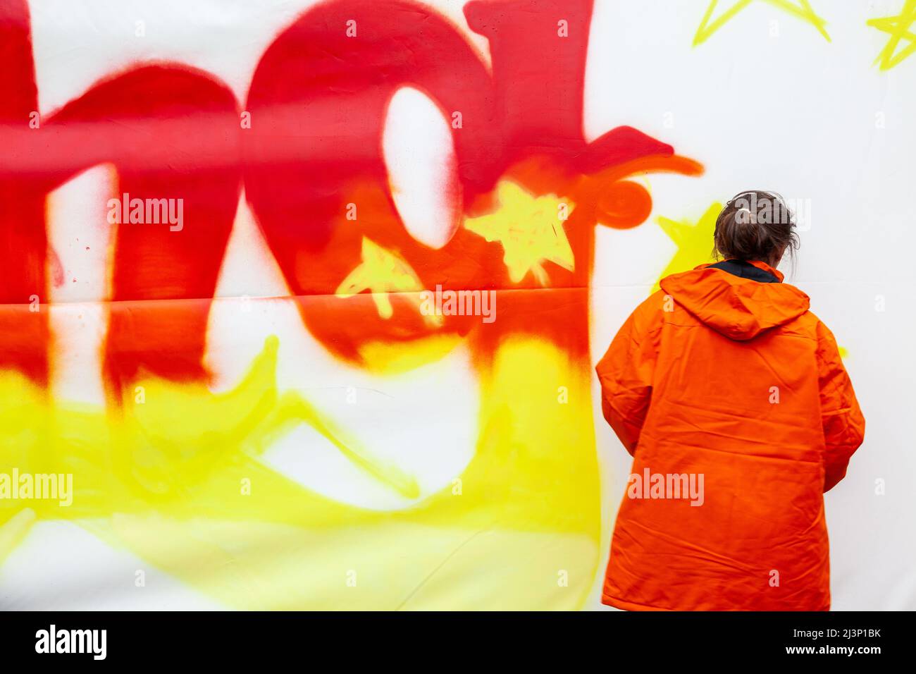 Lady dressed in an orange rain coat tagging a canvas in orange and yellow. Brussels Stock Photo