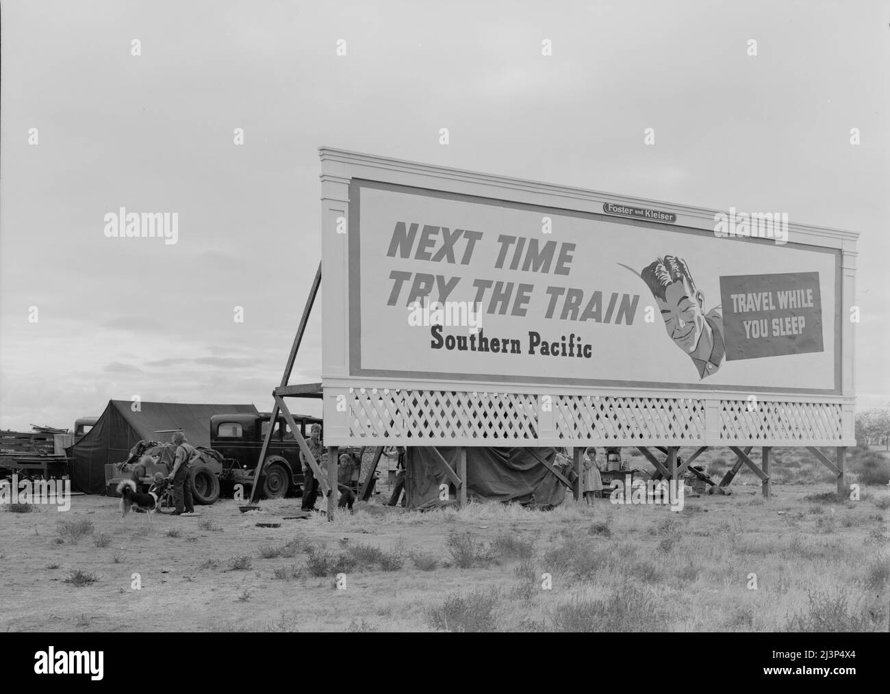 Three families camped on the plains along U.S. 99 in California. They are camped behind a billboard which serves as a partial windbreak. All are in need of work. [Advertisement: 'Next Time Try the Train - Travel While You Sleep - Southern Pacific']. Stock Photo
