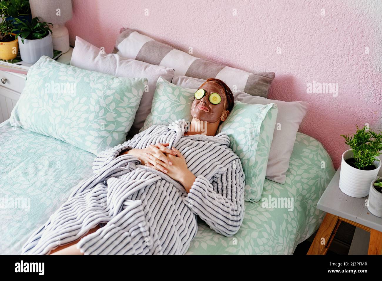 Face mask and chill. High angle shot of an attractive young woman lying on her bed with a mud mask and cucumber slices on her face at home. Stock Photo