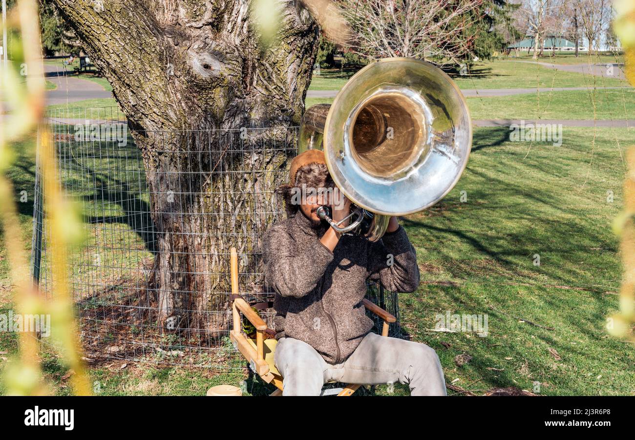 Young man artist with his brown hat plays slow melody on the tuba while sitting on a chair at the park. copy space Stock Photo
