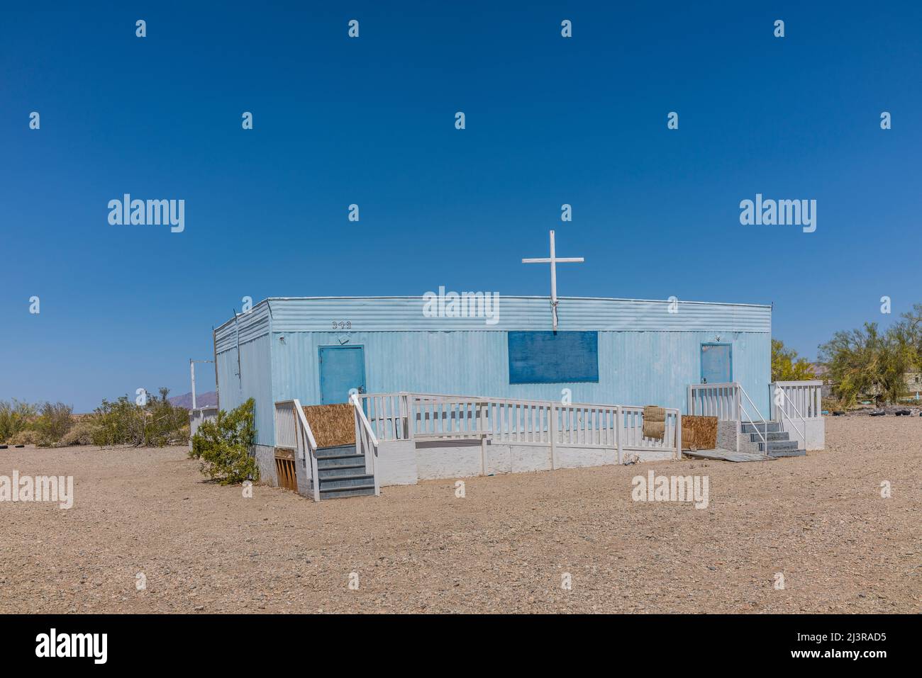 A blue trailer church in Slab City, California with a white wooden cross on top and with stairs on both ends. The church is ADA accessible via a long Stock Photo