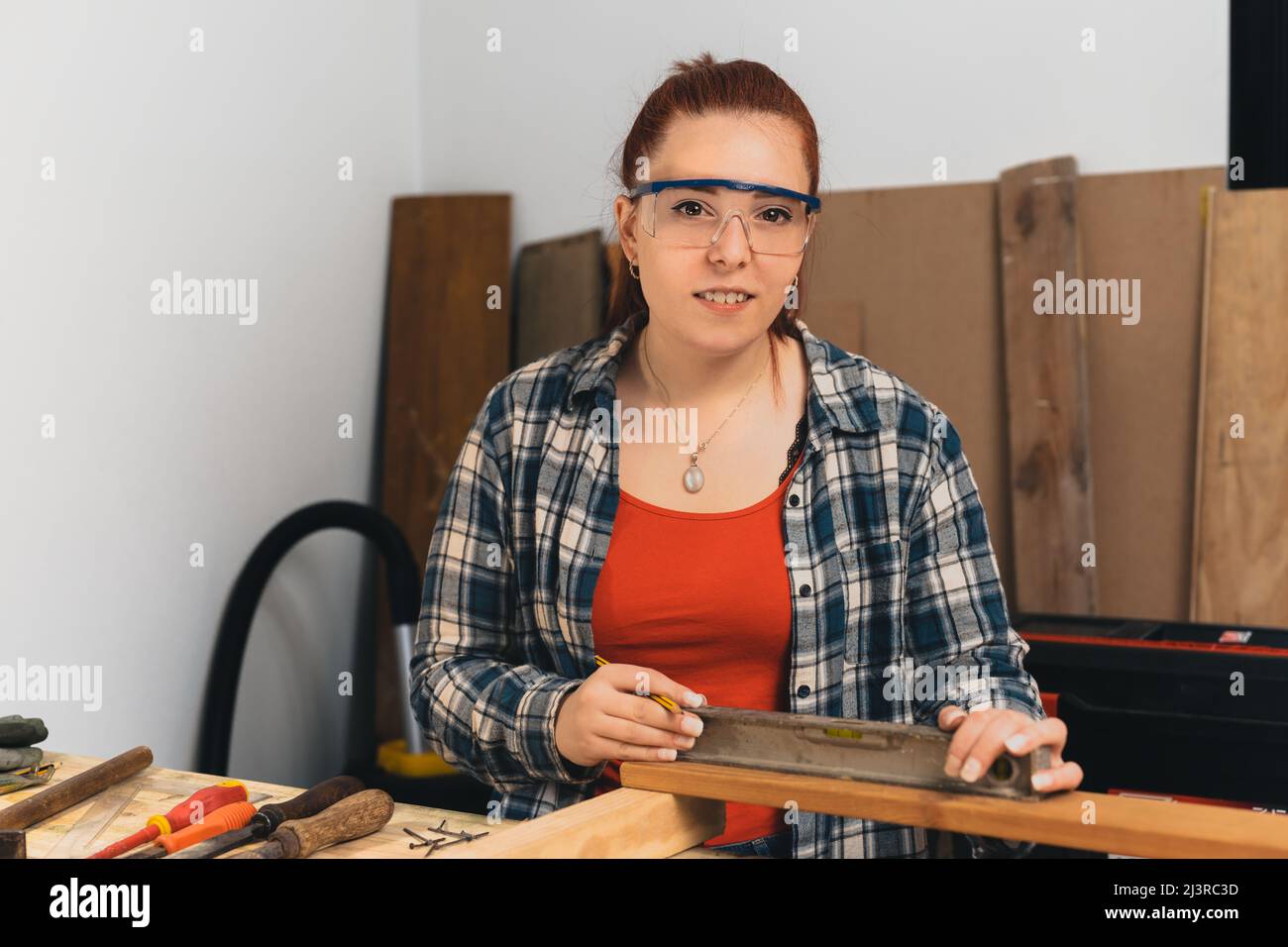 Young red-haired female carpenter using a precision level in her small carpentry workshop. Looking at the camera Stock Photo