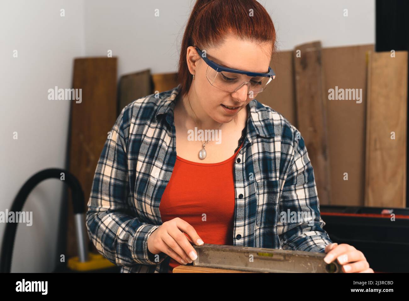 Young red-haired female carpenter using a precision level in her small carpentry workshop. Stock Photo