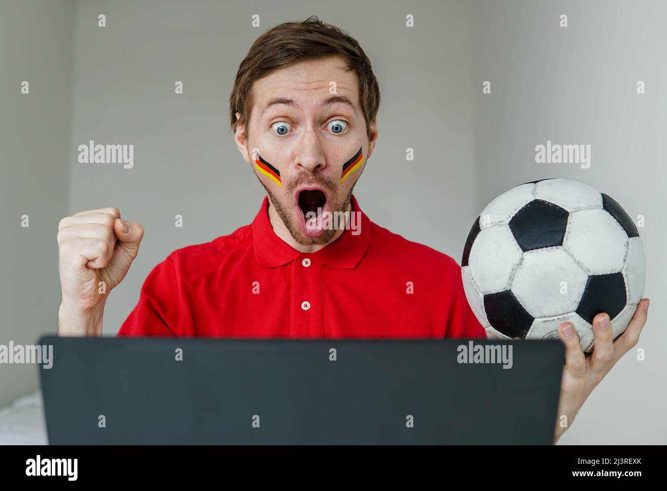Young excited man fan support Germany national football team hold in hand soccer ball watch tv live stream on laptop. Stock Photo