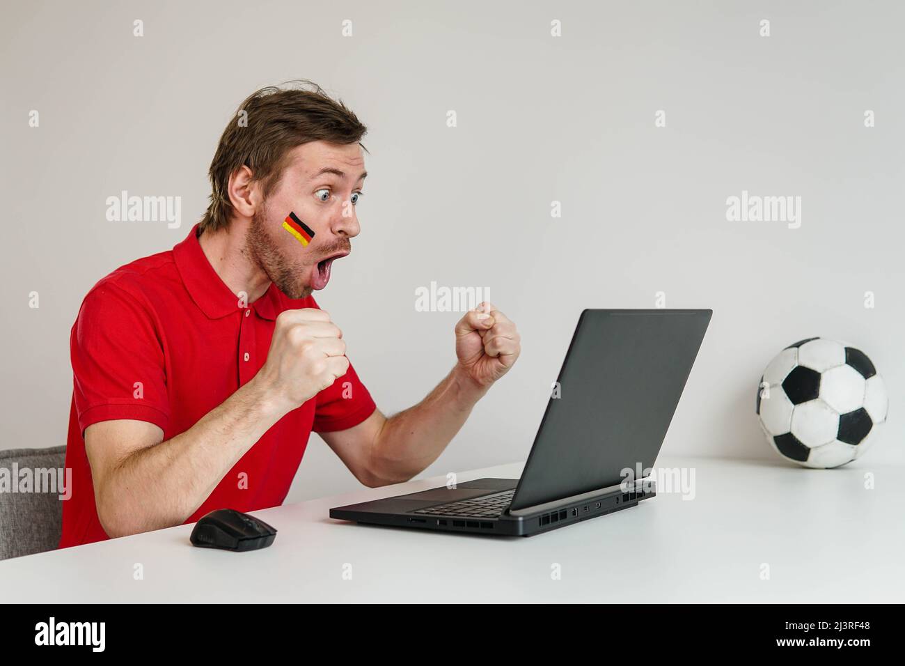 Young excited man fan in red shirt support Germany national football team hold in hand soccer ball watch tv live stream on laptop. Stock Photo