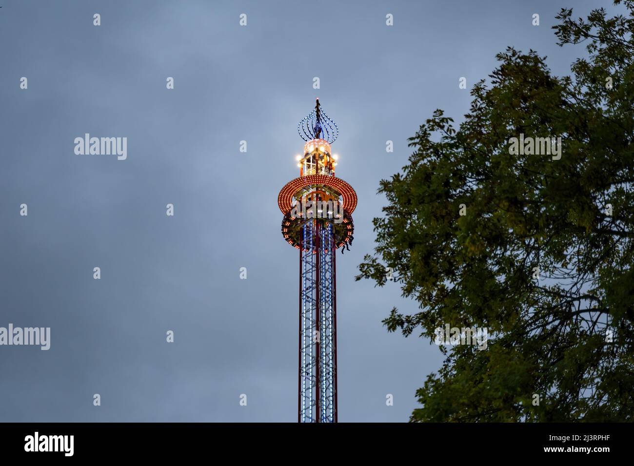 Fairground ride very high in the air in front of a dark sky. The illuminated free fall tower is waiting at the upper position and making people afraid Stock Photo