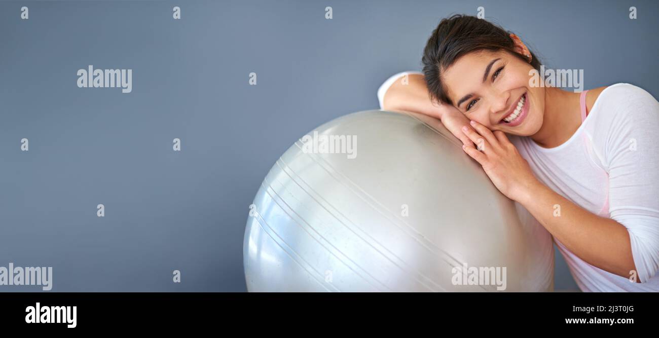 Exercising with my swiss ball makes me happy. Cropped shot of a sporty young woman leaning on a pilates ball against a grey background. Stock Photo