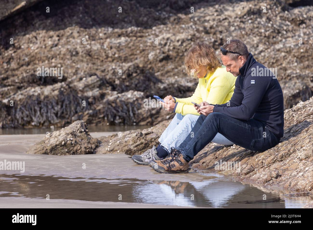 Two people sitting on the rocks at low tide both concentrating on their mobile phones Stock Photo